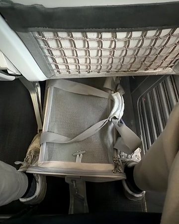 A grey soft-sided cat carrier stowed in the footwell under an aircraft seat, seat pocket net above it